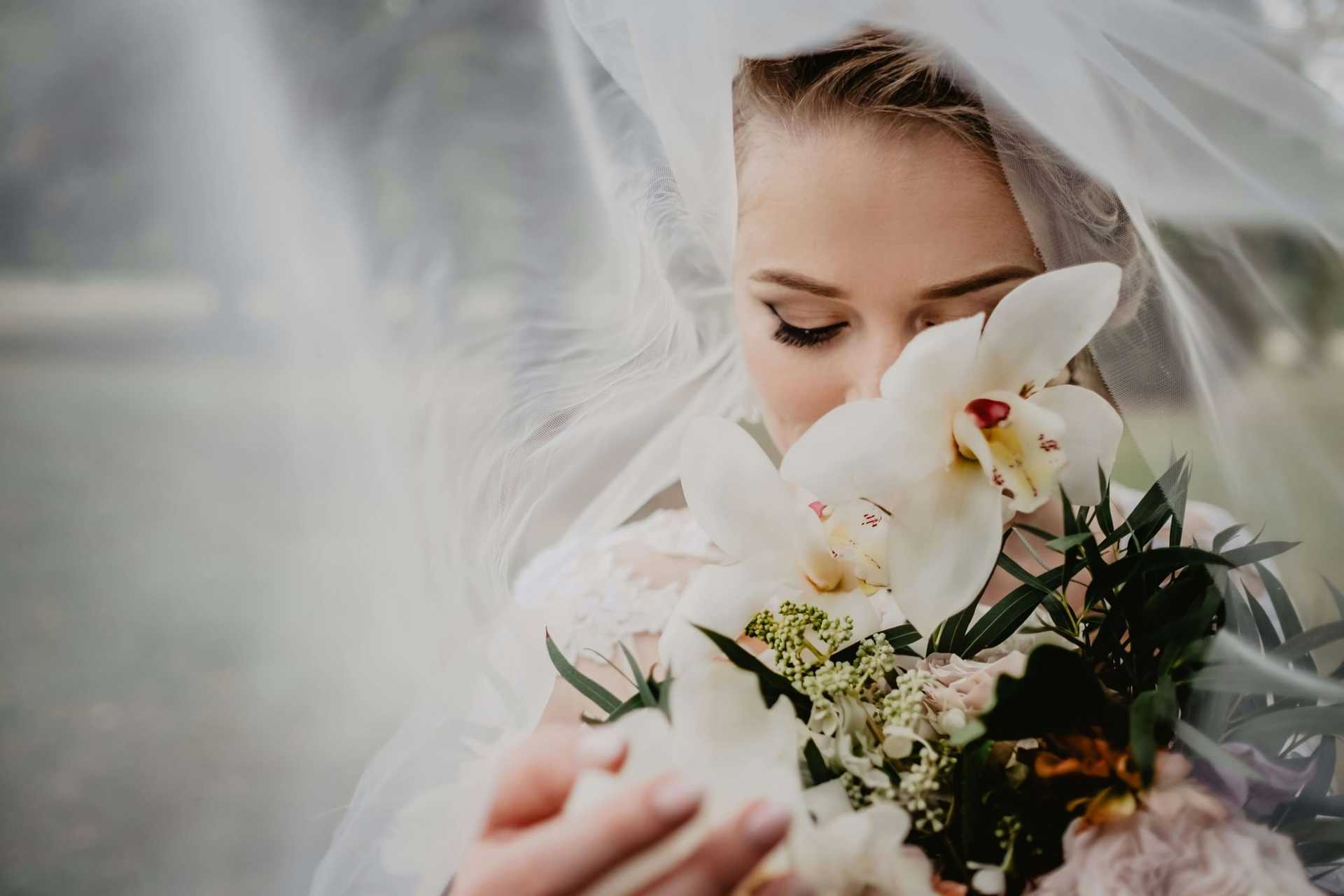Bride with eyes closed, holding orchid bouquet under flowing veil outdoors.