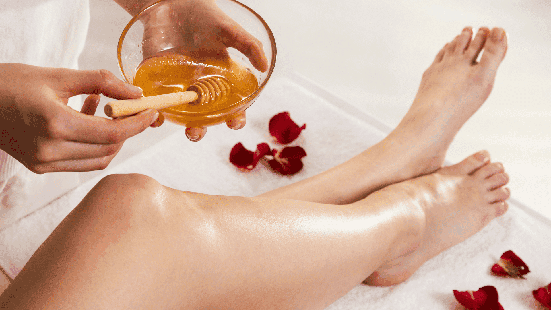 Person applying honey treatment to legs for spa and skincare, surrounded by rose petals.